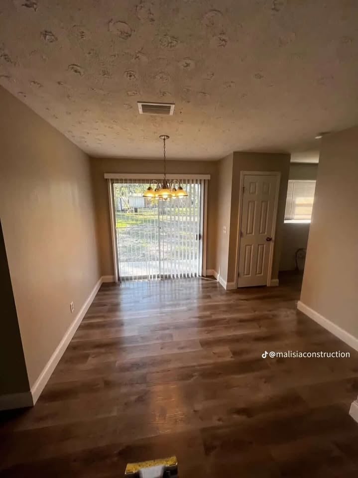 Empty living room with hardwood floors, beige walls, glass sliding doors with blinds, pendant light, and open doorway