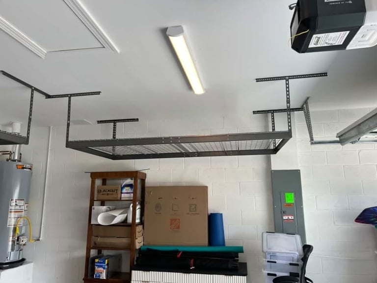 Garage storage area with wall-mounted shelving, overhead storage racks, wooden shelf unit, cardboard boxes, and fluorescent lighting