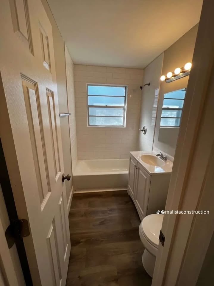 Modern bathroom with white vanity, mirror with lighting, toilet, and white bathtub/shower unit with windows