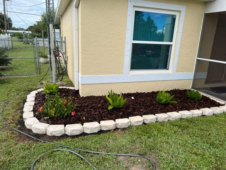 Front garden bed with white stone border, dark mulch, and flowering plants against a beige mobile home with blue window