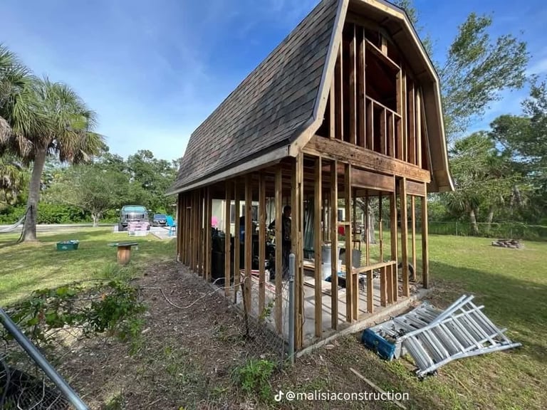 Tiny wooden house under construction with arched roof in grassy outdoor setting surrounded by palm trees and vegetation