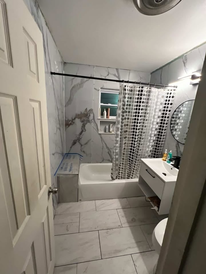Modern bathroom interior with white subway tile flooring, marble-look walls, white vanity sink, round mirror, toilet, and shower with patterned curtain
